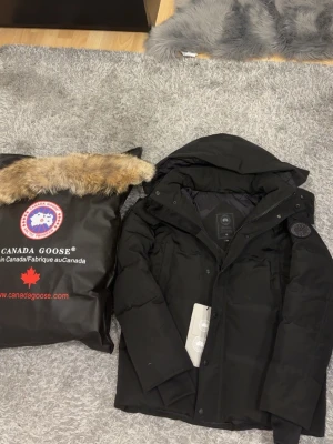 Canada goose black label - Svart dunjacka från Canada Goose med huva och avtagbar pälsdetalj. Jackan har en stilren look med diskret logotyp på ärmen, dragkedja och knappar framtill. Perfekt för kalla vinterdagar och har en klassisk, puffer-design.