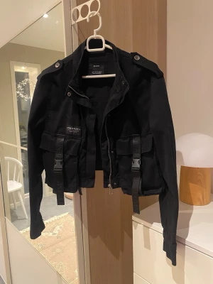Svart croppad jacka från Bershka - Svart croppad jacka från Bershka med långa ärmar, stora fickor framtill och snygga spännen. Jackan har axelklaffar, dragkedja och knappar. Perfekt för dig som gillar streetstyle och vill ha en edgy look.