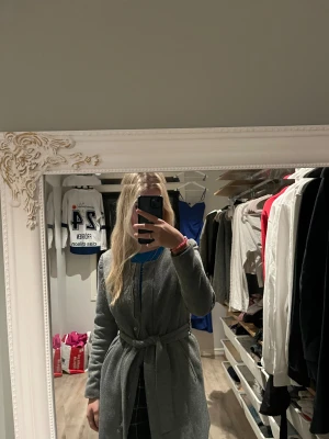 Grå kappa VERO MODA - Säljer en stilren grå kappa från Vero Moda med bälte i midjan och knappar framtill. Jackan har lång ärm, rund hals och är tillverkad i ett mjukt ullmaterial med slät insida. Finns ett hål i tyget i fickan men inget som inte går att laga. Annars är den i jättebra skick.