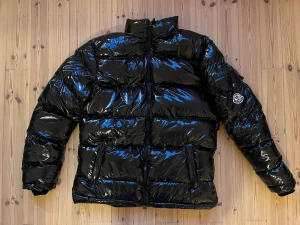 Svart pufferjacka från Moncler - Säljer en glansig svart pufferjacka från Moncler med klassisk logga på ärmen. Jackan har hög krage, dragkedja framtill och stora fickor. Perfekt för kalla dagar och riktigt snygg streetstil. Materialet är syntet och dun, vilket gör den både varm och lätt. Köpt här på Plick, för stor för mig så jag har aldrig använt den.