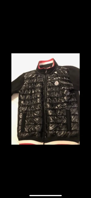 Svart Moncler pufferjacka med detaljer - Snygg svart pufferjacka från Moncler med glansig quiltad front och svarta ärmar. Jackan har dragkedja, Moncler-logga på bröstet och ärmarna samt ribbade muddar med röd och vit rand vid krage och nederkant. Perfekt för dig som vill ha en sportig och trendig look.