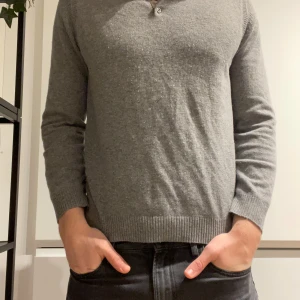 Grå polotröja från Massimo Dutti - Snygg grå polotröja från Massimo Dutti i mjuk ullmix. Tröjan har klassisk krage, två knappar vid halsen och ribbade muddar vid ärmslut och nederkant. Perfekt för dig som gillar stilrena och bekväma plagg.