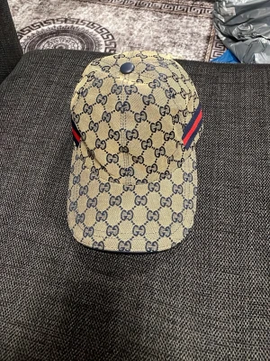 En kaps  - Snygg beige keps från med klassiskt-mönster i mörkblått och bruna detaljer på insidan. Kepsen har justerbar rem baktill och en röd-blå rand på sidan som ger extra lyxig känsla.