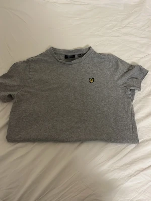 Grå t-shirt från Lyle & Scott - Snygg grå t-shirt från Lyle & Scott med klassisk gul logga på bröstet. T-shirten har rund hals och korta ärmar, tillverkad i mjuk bomull för skön känsla. Perfekt för dig som gillar stilrena och enkla plagg med en touch av märke. Size 14-15, mycket bra skick