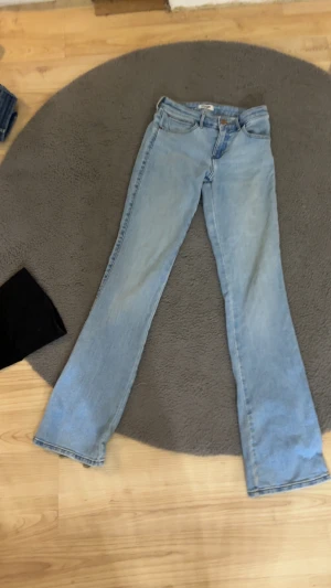 Ljusblå bootcut jeans från Wrangler - Säljer ett par ljusblå jeans från Wrangler med klassiska W-sömmar på bakfickorna. Modellen har bootcut-ben och normal passform. Jeansen är tillverkade i jeansmaterial och har slitningar vid bensluten vilken man ser på bilderna