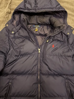 Mörkblå dunjacka från Polo Ralph Lauren - Säljer en mörkblå dunjacka från Polo Ralph Lauren med huva och dragkedja framtill. Jackan har ett rött Polo-logo broderat på bröstet, två sidofickor med dragkedja och quiltad design. Perfekt för kalla dagar och riktigt snygg stil.jackan är i size 18/20 som motsvarar s.skeiv vid frågor. Priset kan diskuteras vid snabba affär.
