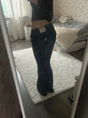 Mörkblå bootcut jeans True Religion - Säljer ett par mörkblå jeans från True Religion med bootcut passform och låg midja. Jeansen har klassiska fickor med dekorativa sömmar och silverfärgade knappar. Materialet är jeans och de har en snygg, figurnära siluett med utsvängda ben. Storlek W27!