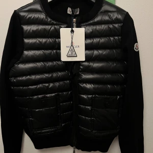 Svart quiltad cardigan jacka Moncler - Svart cardigan jacka från Moncler med quiltad, glansig framsida i syntet och två stora fickor med knappar. Ärmar och rygg är stickade för en snygg kontrast. Klassisk rund halsringning och dragkedja framtill. Perfekt för dig som gillar stilren och sportig look.