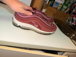 Nike Air Max 97 vinröd/lila sneakers - Nike Air Max 97 sneakers i vinrött och lila med vågiga linjer och vit sula med synlig Air-enhet. Skorna har snörning och en ikonisk, sportig design. Materialmix av syntet och textil. Perfekta för dig som vill sticka ut med färg och stil.