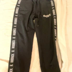 Svarta Hoodrich mjukisbyxor med tryck - Svarta mjukisbyxor från Hoodrich med vit logotyp längs båda benen och på ena fickan. Byxorna har snörning i midjan och är tillverkade i mjukt material som passar chill och streetwear. Snyggt tryck ger en modern vibe.