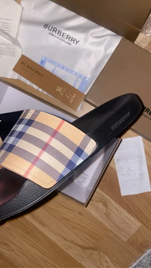 Burberry sandaler med klassiskt rutmönster - Snygga sandaler från Burberry med den ikoniska rutan i beige, blått och rött på remmen. Svart platt sula med Burberry-logga på sidan och undersidan. Perfekt för dig som vill ha en lyxig och stilren look i sommar. Materialet är syntet och skinnliknande.