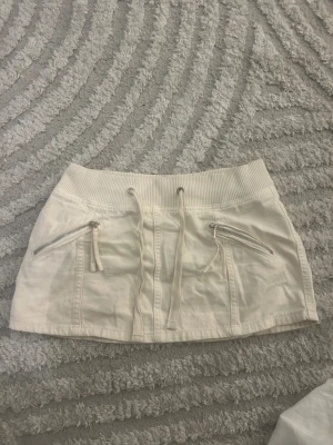 Vit kortkjol från Pull&Bear med snörning - Snygg vit kortkjol från Pull&Bear i bomull med bred ribbad midja och snörning framtill. Kjolen har två dragkedjefickor på framsidan. Säljer för att jag aldrig får tillfälle att använda den.