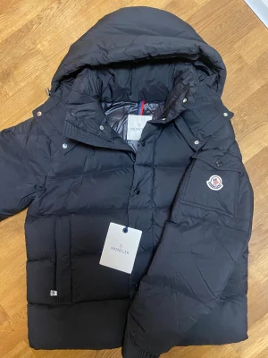 Svart Moncler pufferjacka - Säljer en svart pufferjacka från Moncler med stor huva och feta, quiltade sektioner. Jackan har Moncler-logga på ärmen, praktiska fickor och både knappar och dragkedja framtill. Perfekt för dig som vill ha en stilren och varm jacka. Jackan är perfekt till vintern. 10 utav 10 en super fet och unik jacka. 