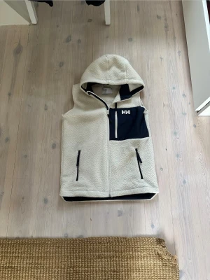 Helly Hansen väst - Säljer en beige fleeceväst från Helly Hansen med huva och svart bröstficka med HH-logga. Västen har dragkedja framtill och två sidofickor med svarta detaljer. Perfekt för lager-på-lager och chill dagar utomhus.