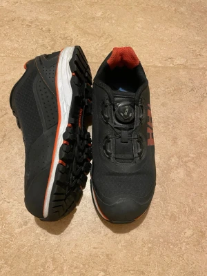 Svarta sneakers med BOA-snörning - Säljer ett par svarta sneakers från Helly Hansen med röd detalj och vit sula. Skorna har grov yttersula för extra grepp och BOA-snörningssystem för smidig justering. Ovandelen är i syntetmaterial och textil, perfekt för dig som gillar att vara aktiv.