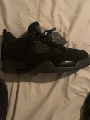 Svarta Nike Air Jordan 4 sneakers - Nike Air Jordan 4 sneakers i helsvart färg med klassisk Jumpman-logga på plösen och innersulan. Skorna har snörning, meshpaneler på sidorna och detaljer i syntet och skinn. Snygg, sportig siluett med platt sula och rund tå. Perfekt för dig som gillar streetwear.