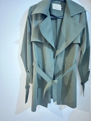 Grön trenchcoat från Coco Miss - Snygg grön trenchcoat från Coco Miss med bred krage, knytbälte i midjan och detaljer vid ärmsluten. Jackan har en stilren look och är tillverkad i ett mjukt syntetmaterial. Perfekt för dig som gillar klassisk stil med modern touch.