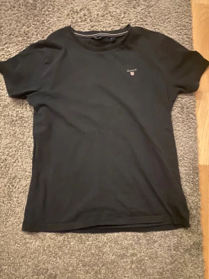 Navy Blå t-shirt från GANT med logga - Navy blå t-shirt från GANT med klassisk rund hals och liten broderad logga på bröstet. Tillverkad i mjuk bomull som känns skön mot huden. Enkel och stilren design som funkar till det mesta. Perfekt för dig som gillar cleana plagg med diskret branding.