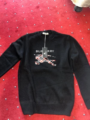 Svart stickad Burberry tröja - Svart stickad tröja från Burberry med klassiskt Burberry-tryck och broderad logga på bröstet. Tröjan har rund hals, ribbade muddar och långärmad passform. Detaljer i vitt och rött mönster på framsidan ger en snygg kontrast. Perfekt för dig som gillar stilrena märkesplagg.