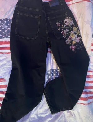 Svarta jeans med broderade blommor - Unika svarta jeans med snyggt blommigt broderi på ena bakfickan och benet. Jeansen har kontrastsömmar och klassisk femficksdesign. Passformen är rak och materialet är robust denim. Perfekt för dig som vill sticka ut med coola detaljer.