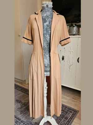 Beige långklänning/kofta - Snygg beige långklänning/kofta, med korta ärmar och svarta kontrastkanter vid ärmslut och fickor. Knytband i midjan för extra form. Tillverkad i mjukt, tunt material. 