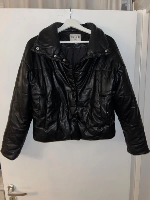 Svart pufferjacka i skinn från NA-KD - Säljer en svart pufferjacka från NA-KD i storlek 36. Jackan har en cool oversized look, bred krage med tryckknappar och två fickor framtill. Materialet är syntetiskt skinn som ger en snygg glansig finish. Perfekt för dig som vill ha en trendig och varm jacka.
