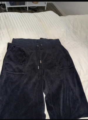 Svarta mjukisbyxor Juicy Couture M - Svarta mjukisbyxor från Juicy Couture i storlek M. Byxorna har bred resår i midjan med snörning, raka ben och stora fickor fram samt en bakficka med knappar. Mjukt och glansigt material som känns lyxigt och skönt.