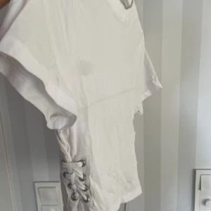 Vit t-shirt med snörning i sidan - Snygg vit t-shirt med korta ärmar och cool snörningsdetalj i sidan. Modellen är enkel men har en twist med metallöljetter och snören som ger en trendig vibe. Perfekt till jeans eller shorts för en chill look. Materialet känns mjukt och skönt mot huden.