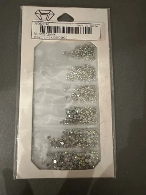 Strass-stenar för nageldekoration - Säljer ett paket med strass-stenar i olika storlekar och former, perfekta för att dekorera naglarna. Stenarna är silvriga och skimrande, och kommer i en praktisk plastförpackning med flera fack. Materialet är plast med glittrande yta.