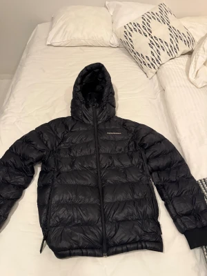 Svart dunjacka från Peak Performance - Säljer en svart dunjacka från Peak Performance med huva och dragkedja framtill. Jackan har quiltad design och är perfekt för kalla dagar. Den är lätt och har en snygg, sportig look med diskret logga på bröstet. Passar dig som gillar stilren och funktionell ytterjacka.