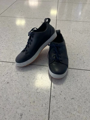 Mörkblå sneakers med vit sula - Snygga mörkblå sneakers med rund tå och vit platt sula. Skorna har snörning och är tillverkade i ett slätt, lätt texturerat skinnmaterial. Perfekt för dig som gillar stilrena och klassiska sneakers med en modern touch.