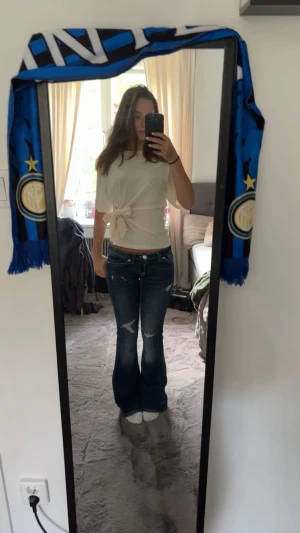 Blå bootcut jeans med slitningar - Säljer ett par mörkblå bootcut jeans med snygga slitningar på knäna och ljusare tvätt på låren. Jeansen har låg midja. 