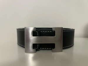 Svart bälte  med H-spänne i metall - Stilrent svart skärp med grov struktur och vit kontrastsöm längs kanterna. Skärpet har ett stort, silvrigt H-format metallspänne som ger en lyxig vibe. Perfekt för att lyfta din outfit med en snygg detalj. Passar till jeans eller kostymbyxor.storlek 46/115 går att ändra. Lägg gärna ett bud, jag är ganska lätt att pruta ner