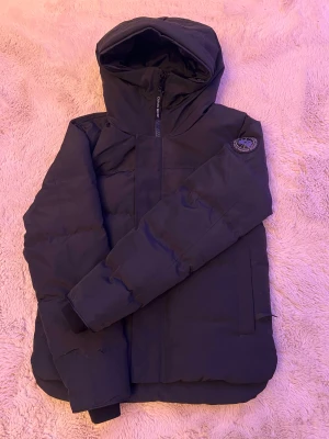Svart pufferjacka från Canada Goose - Säljer en svart pufferjacka från Canada Goose med huva och diskret logga på ärmen. Jackan har dragkedja framtill, två stora fickor och är perfekt för kalla dagar. Klassisk, stilren design och riktigt varm. Levereras med originalväska.