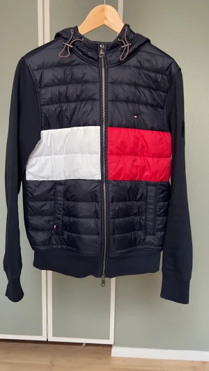 Tommy Hilfiger pufferjacka, cardigan med färgblock - Snygg pufferjacka från Tommy Hilfiger med klassiska färgblock i svart, vitt och rött. Jackan har dragkedja framtill, huva med snörning och ribbade muddar. Ikonisk logga på ärmen och quiltad front. Perfekt för dig som vill sticka ut med stil. Storlek M passar S även, nyskick använd ett par gånger.