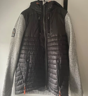 Svart cardigan/puffer från Superdry - Säljer en svart pufferjacka från Superdry med gråa ärmar och huva. Jackan har quiltad framsida och rygg, två dragkedjefickor med orange detaljer och Superdry-logga på axeln. Perfekt för lite mindre kalla dagar nu under vintern.😁bvsa