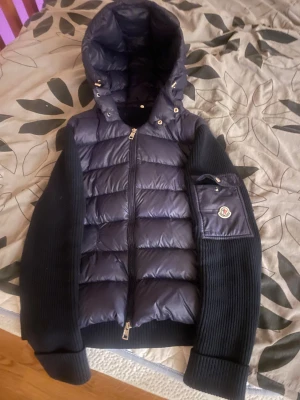 Svart Moncler pufferjacka med huva - Svart pufferjacka från Moncler med stor huva och ribbstickade ärmar. Jackan har en quiltad front, dragkedja och en ficka med Moncler-logga på ärmen. Materialmix av polyester och stickad ull ger både värme och stil. Perfekt för kyliga dagar. Det har funnits ett litet hål framför jackan men det är sytt så inget problem alls.Priset är 2200kr,men om det nog är för mycket så kan vi lösa ett bättre pris privat. 