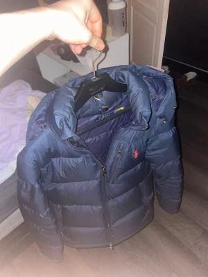 Mörkblå dunjacka Polo Ralph Lauren XS - Snygg mörkblå dunjacka från Polo Ralph Lauren i storlek XS. Jackan har huva, två sidofickor med dragkedja och klassisk röd Polo-logga på bröstet. Materialet är glansigt och quiltat, perfekt för kalla dagar. Performance-modell med hög krage och dragkedja framtill.