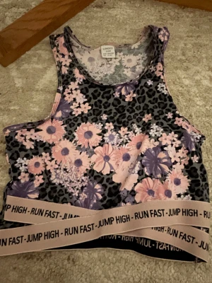 Blommig leopardmönstrad linnetopp Lindex - Cool linnetopp från Lindex med svart leopardmönster och blommor i rosa och lila. Elastiskt band nedtill med texten 'RUN FAST - JUMP HIGH' i svart. Perfekt för träning eller sportiga dagar. Materialet är mjukt och stretchigt polyester.