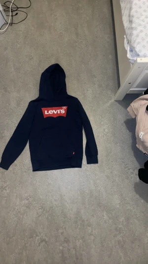 Blå hoodie från märket Levi’s - En klassisk blå hoodie från Levi’s, tillverkad i ett mjukt och bekvämt bomullsmaterial. Hoodien har en tidlös design med den ikoniska röda Levi’s-loggan placerad mitt på bröstet, vilket ger ett tydligt och stilrent uttryck. Perfekt för vardagsbruk och lätt att matcha med både jeans och joggers.