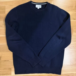 Marinblå stickad tröja från Lacoste - Snygg marinblå stickad tröja från Lacoste med diskret logga på bröstet. Tröjan har rund hals, ribbade muddar och subtila vertikala ränder i stickningen. Perfekt för dig som gillar klassisk och stilren design.