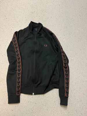 Svart Fred Perry bomberjacka S - Svart sip/hoodi från Fred Perry i storlek S. Sipen har röda detaljer längs ärmarna med Fred Perry-loggor, hel dragkedja framtill och ribbade muddar. Materialet är mjukt och syntetiskt, perfekt för en sportig och clean look.