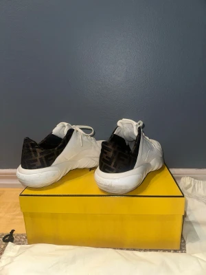 Fendi vita sneakers med FF-logga - Säljer ett par vita Fendi sneakers med svart FF-logga på hälen. Skorna har snörning, rund tå och är tillverkade i en mix av skinn och textil. Snygg chunky sula och klassisk Fendi-detalj baktill. Kommer med originalkartong och dustbags.