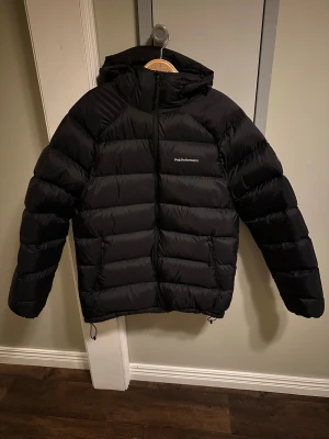 Svart dunjacka Peak Performance XL - Svart dunjacka från Peak Performance i storlek XL. Jackan har huva, dragkedja framtill och två sidofickor med dragkedja. Klassisk pufferstil med broderad logga på bröstet och ärmen. Perfekt för kalla dagar när du vill hålla värmen och ändå se clean ut.