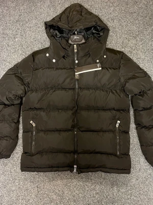 Svart pufferjacka med huva från Stone Island - Snygg svart pufferjacka från Stone Island med huva och emblem på ärmen. Jackan har flera dragkedjor, quiltad design och praktiska fickor. Perfekt för kalla dagar och har en modern, loose passform som ger en cool look.