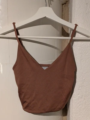 Brun croppad linnetopp från Missguided - Säljer en brun croppad linnetopp från Missguided med smala axelband och v-ringad hals. Toppen har en enkel och stilren design som är perfekt för att skapa en chill och trendig look. Materialet är mjukt och lätt, vilket gör den skön att bära.