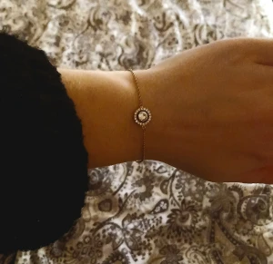 Guldigt armband med glittrande sten - Snyggt och tunt armband i guldfärgad metall med en rund, gnistrande sten i mitten omgiven av små kristaller. Lätt kedja och justerbar längd med karbinlås. Perfekt för dig som gillar stilrena smycken med lite extra bling.