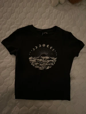 Svart t-shirt med månfaser och vågor - Cool svart t-shirt från H&M med ett stort tryck framtill som visar månfaser och vågor i vitt. Rund hals och korta ärmar, tillverkad i mjuk bomull. Passformen är normal och trycket ger en unik vibe till din outfit.
