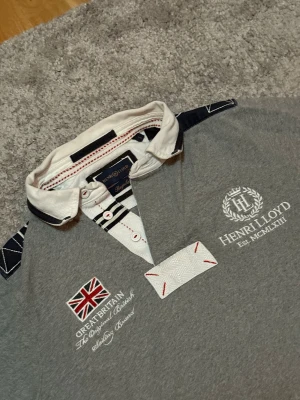 Henri Lloyd pikétröja grå  - Säljer en grå långärmad pikétröja från Henri Lloyd i storlek S, med vit krage och snygga broderade detaljer. Tröjan har brittisk flagga, logga och text framtill, samt kontrastpaneler på axlar och ärmar. Knappar vid halsen och coola sömmar i rött och vitt. Perfekt för dig som gillar sportig stil.