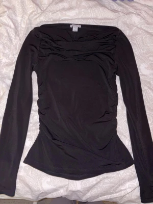 Svart långärmad topp från Gina Tricot - Säljer en svart långärmad topp från Gina Tricot med snygga rynkade detaljer vid bröstet och sidorna. Toppen har en slim passform och är tillverkad i mjukt, stretchigt syntetmaterial. Perfekt för dig som gillar stilrena och enkla plagg med en twist.